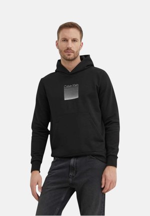 Mand iført sort Calvin Klein hoodie med frontlomme og mørke jeans, stående mod en ensfarvet baggrund.