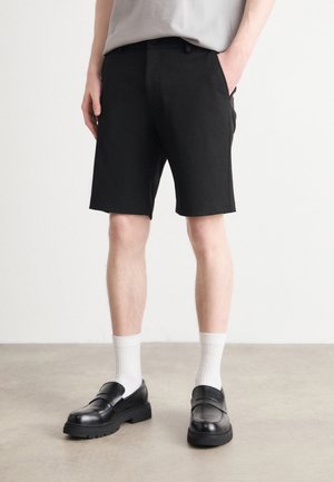Pantalones cortos negros con un ajuste a medida, que presentan bolsillos laterales, combinados con calcetines blancos y mocasines negros. Tejido suave, diseño minimalista.