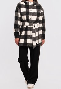 Černobílá kostkovaná fleecová bunda s kapucí, předními knoflíky a látkovým páskem, kombinovaná s černými kalhotami a bílými teniskami.