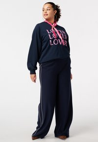 Navy sweatshirt met roze "LOVER" tekst, gecombineerd met wijde navy broek met witte zijstrips. Model draagt een roze sjaal en schoenen met dierenprint.