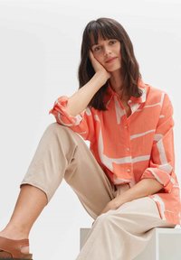 Korallenfarbenes Hemd mit weißen geometrischen Mustern, hochgekrempelten Ärmeln, kombiniert mit beigen Hosen und braunen Sandalen, in Sitzpose.