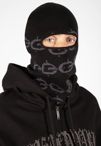 Pasamontañas negro de punto con un patrón circular gris y dos aberturas para los ojos; combinado con una sudadera con cremallera negra a juego.