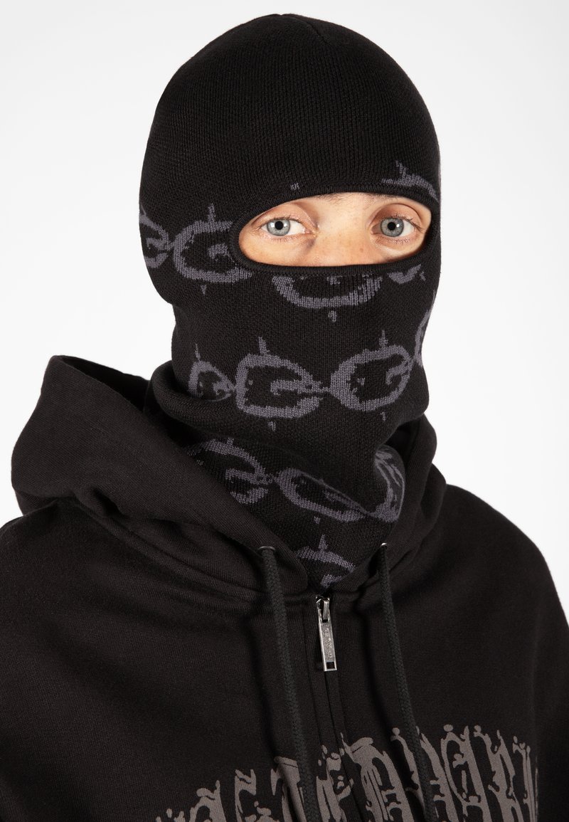 Pasamontañas negro de punto con un patrón circular gris y dos aberturas para los ojos; combinado con una sudadera con cremallera negra a juego.
