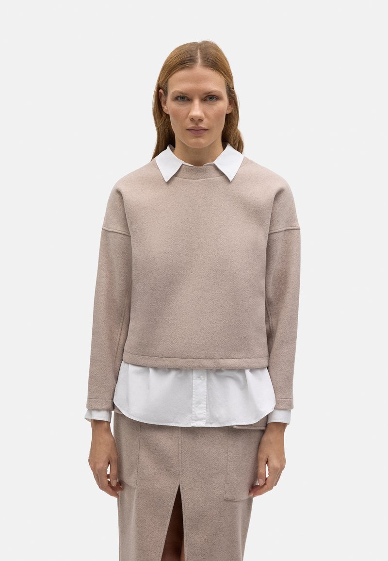 Suéter corto color beige claro con un acabado texturizado, superpuesto a una camisa blanca clásica con cuello, combinado con una falda a juego que presenta una abertura.