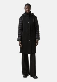 Sort frakke med pufærmer, frontknapper, to store lommer og hætte. Glat stof med et kontrasterende skinnende materiale på ærmerne.