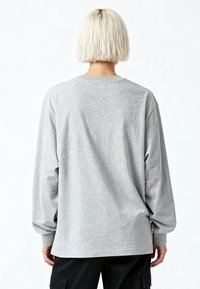 Person mit kurzen blonden Haaren, die ein hellgraues Langarmshirt und schwarze Hosen trägt, steht mit dem Rücken zur Kamera vor einem weißen Hintergrund.