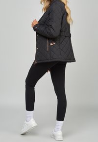 Chaqueta negra acolchada con herrajes en color oro rosa, cintura con cinturón y bolsillos con cremallera en los laterales, combinada con leggings negros y zapatillas blancas.