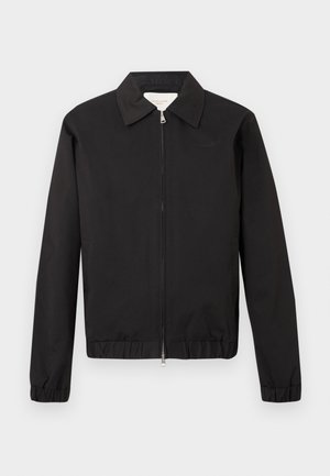 Geacă neagră cu fermoar, guler, manșete și tiv elastice, cu un logo subtil brodat pe pieptul stâng, afișată pe un fundal simplu.