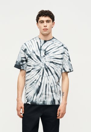 Νέος άνδρας με κοντά σγουρά μαλλιά, φορώντας μαύρο-άσπρο tie-dye κοντομάνικο μπλουζάκι και μαύρο παντελόνι, στέκεται μπροστά σε απλό ανοιχτό φόντο.