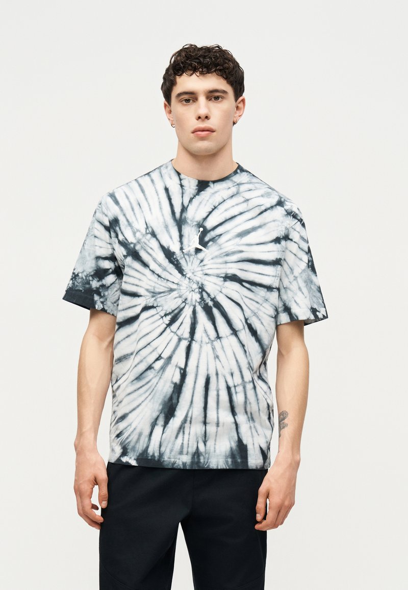 Νέος άνδρας με κοντά σγουρά μαλλιά, φορώντας μαύρο-άσπρο tie-dye κοντομάνικο μπλουζάκι και μαύρο παντελόνι, στέκεται μπροστά σε απλό ανοιχτό φόντο.