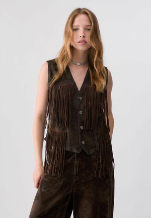 Kobieta z długimi falującymi włosami ma na sobie ciemną, bezrękawną kamizelkę z frędzlami i guzikami, pasujące ciemne spodnie oraz naszyjnik typu choker, stojąc na tle prostego tła.