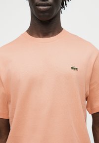 Homme portant une chemise à manches courtes couleur pêche pâle avec un petit logo de crocodile vert sur le côté gauche de la poitrine, sur un fond uni.