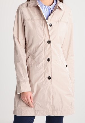 Femme portant un manteau léger beige boutonné sur une chemise rayée bleue et blanche à col.