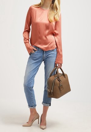 Top a maniche lunghe in satin rosa, jeans azzurri chiari, tacchi nudi e una borsa in pelle marrone con cerniere e un design a forma di etichetta di bagaglio.