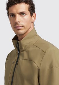 Lichte khaki jas met hoge kraag, ritssluiting en textuurstof. Heeft een schoudernaden detail en een logo op de mouw.