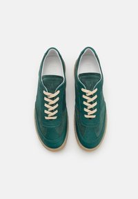 Pár zelených kožených a semišových tenisek se béžovými tkaničkami a gumovými podrážkami, vystavených z vrchu na bílém pozadí.