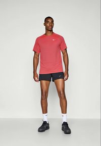 Piros, rövid ujjú sportpóló texturált anyagból, Nike logóval, fekete atlétikai rövidnadrággal és fekete sportcipővel párosítva. Fehér zoknik láthatók.