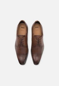 Barna bőrből készült dress shoes, texturált felülettel, hegyes orral és négy fűzőlyukkal. Diszkrét, kontrasztos öltés részletekkel rendelkezik.