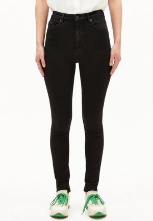 Femme portant un jean skinny noir taille haute associé à des baskets blanches et vertes, debout devant un fond blanc.