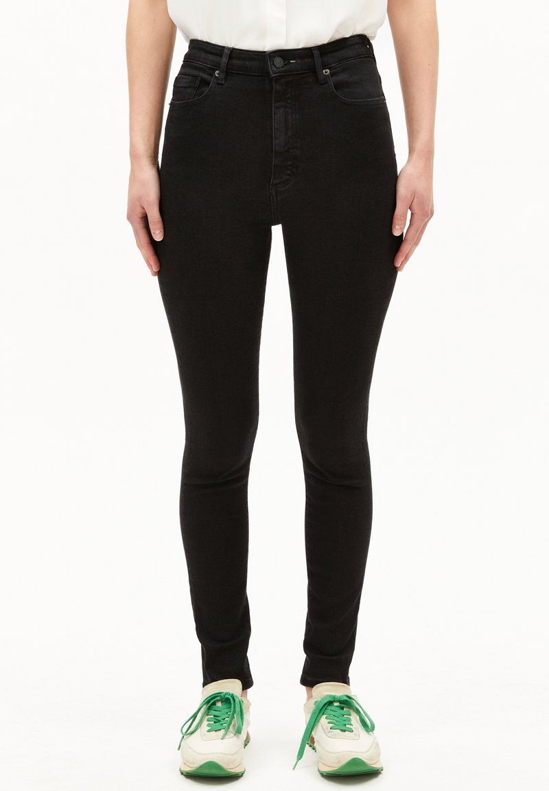 Femme portant un jean skinny noir taille haute associé à des baskets blanches et vertes, debout devant un fond blanc.