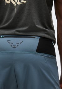 Κοντινό πλάνο ενός ατόμου που φοράει μπλε αθλητικό παντελόνι με τσεπάκι με φερμουάρ και μαύρο λογότυπο γάτας στη ζώνη κάτω από σκούρο γκρι πουκάμισο.