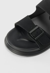 Svart sandal laget av mykt materiale, med to brede stropper med rektangulær spenne, og en avrundet såle med en glatt overflate.
