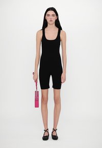 Bodysuit noir sans manches avec encolure arrondie et design de short de vélo. Le modèle tient un petit sac à main rose vif. Chaussures noires à pois.