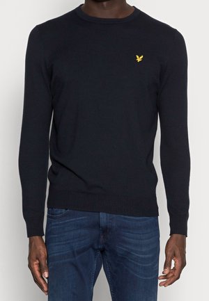 Mand iført en tætsiddende sort langærmet striktrøje med et lille gult fuglelogo på brystet og mørkeblå jeans.