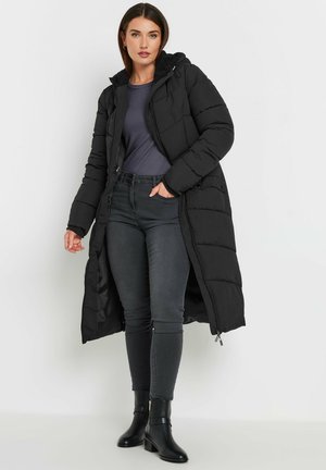 Cappotto lungo imbottito nero con cappuccio, dotato di chiusura con zip e due tasche frontali. Indossato sopra un top scuro, abbinato a jeans attillati e stivaletti.