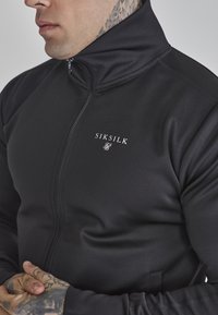 Chaqueta negra con cremallera hecha de un material suave y elástico. Presenta un cuello alto y un pequeño logo blanco en el lado izquierdo del pecho.
