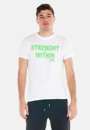 Jeune homme portant un t-shirt blanc avec un texte vert "STRENGHT IS WITHIN" et un pantalon noir, debout devant un fond uni.