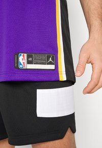 Maglia sportiva viola con pantaloni corti neri. La maglia presenta accenti gialli e un'etichetta con il logo NBA. I pantaloni corti hanno una patch bianca testurizzata.