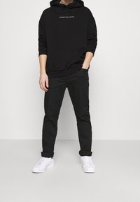 Sudadera de algodón negra con bolsillo frontal y texto blanco "CONSCIOUS MIND", combinada con pantalones negros y zapatillas blancas.