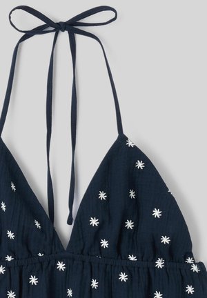 Haut dos nu bleu marine avec des motifs d'étoiles blanches brodées et une lanière nouée autour du cou sur un fond clair uni.