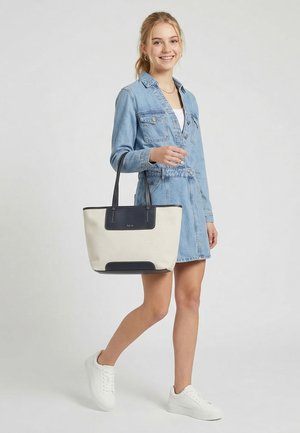 Young woman smiling, wearing a light denim dress, white sneakers, and carrying a beige tote bag with black handles and accents.