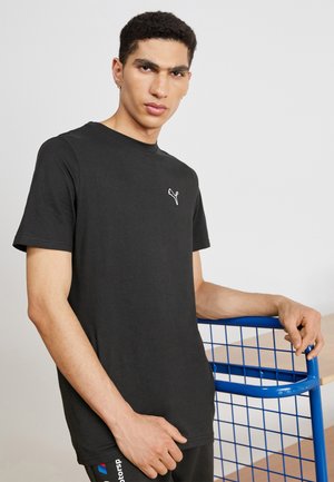 Camiseta de algodón negra con cuello redondo y mangas cortas, que presenta un logo blanco en el pecho. El modelo está de pie contra una estructura de rejilla azul.