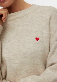 Primo piano di una persona che indossa un maglione lavorato a maglia beige con un piccolo cuore rosso ricamato sul petto, mano poggiata vicino al mento.