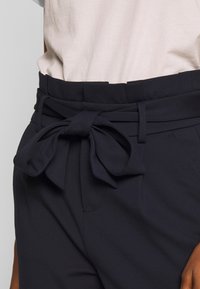 Pantalon bleu marine avec une large ceinture et un grand nœud décoratif à l'avant, fabriqué dans un tissu doux et texturé.