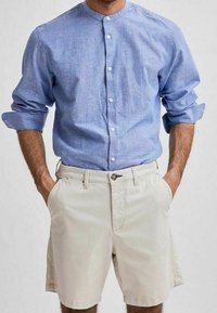Camicia azzurra a righe con colletto alla mandarino, abbinata a pantaloni corti beige con chiusura a bottone singolo e tasche laterali.
