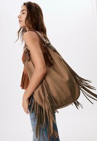 Braune Wildleder-Schultertasche mit langen Fransen, runder Form und verstellbaren Tragriemen. Glatte Textur und dezente goldfarbene Hardware-Akzente.