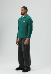 Turkooise fleece trui met een gestructureerd oppervlak, kraag en geborduurde details. Gecombineerd met zwarte wijde broekspijpen en chunky zwarte schoenen.