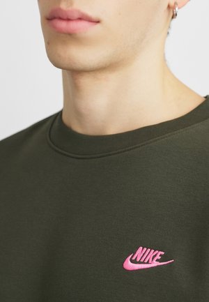 Zbliżenie osoby noszącej oliwkowo-zieloną koszulę z małym różowym logo Nike na lewej piersi oraz srebrną kolczyk kołową.
