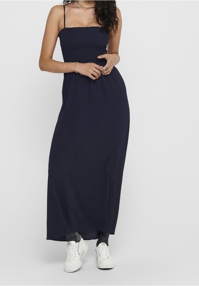 Jeune femme portant une robe maxi noire sans manches à fines bretelles, des baskets blanches et des chaussettes courtes foncées, debout devant un fond uni.
