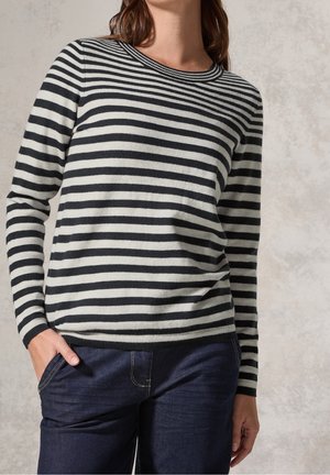 Femme portant un pull rayé noir et blanc à manches longues et un jean bleu foncé avec une main dans la poche.