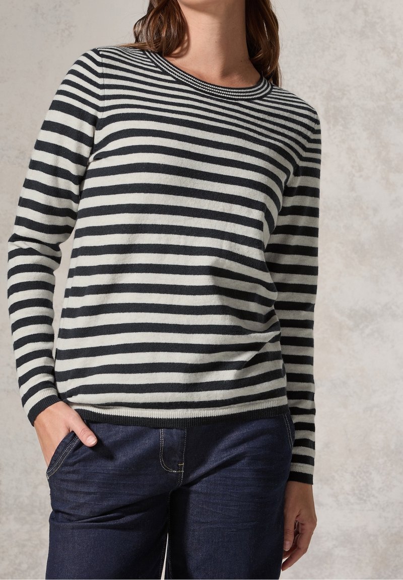 Femme portant un pull rayé noir et blanc à manches longues et un jean bleu foncé avec une main dans la poche.