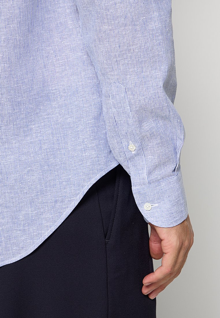 Primer plano de la mano de una persona descansando junto a unos pantalones azul marino, vistiendo una camisa de manga larga a rayas azul claro y blanca con puño abotonado.