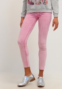 Henkilö, jolla on päällä vaaleanpunaiset skinny-farkut, harmaa kukkakuviollinen collegepaita ja kiiltävät hopeiset nauhalliset kengät matalilla puukoroilla.