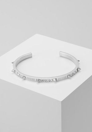 Bracelet manchette ouvert en argent avec des pointes et des cristaux clairs, gravé du mot « GUESS », présenté sur une surface blanche inclinée.
