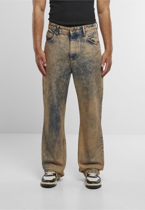 Homme portant un jean ample délavé beige et bleu à effet déchiré, avec des baskets noires et blanches, debout devant un fond uni.