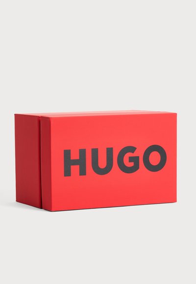 Une boîte HUGO rectangulaire rouge vif, ornée de grandes lettres noires, sur un fond gris clair.